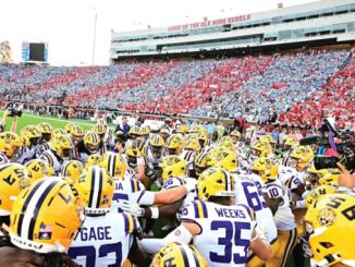 LSU Tigers football players in yellow helmets huddle on the field before kickoff, stadium full of red and blue-clad fans in the stands.