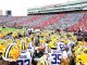 LSU Tigers football players in yellow helmets huddle on the field before kickoff, stadium full of red and blue-clad fans in the stands.
