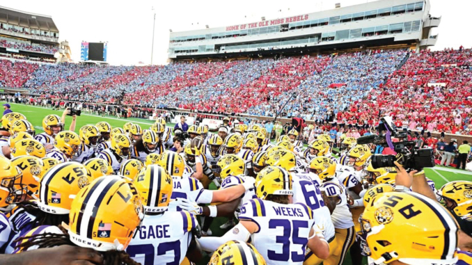 LSU Tigers football players in yellow helmets huddle on the field before kickoff, stadium full of red and blue-clad fans in the stands.