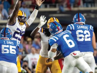 American football players in blue and white uniforms tackle a yellow-uniform opponent on a crowded stadium field.