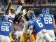 American football players in blue and white uniforms tackle a yellow-uniform opponent on a crowded stadium field.