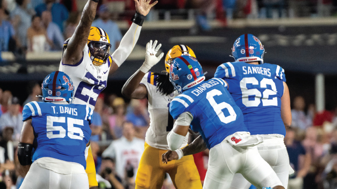 American football players in blue and white uniforms tackle a yellow-uniform opponent on a crowded stadium field.