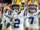 Two LSU football players celebrate on the field, white uniforms with purple numbers 2 and 7 and yellow helmets.