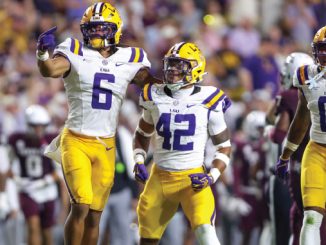 LSU Tigers football players in white jerseys and yellow pants celebrate on the field, with #6 pointing and #42 standing beside, as other teammates celebrate nearby.