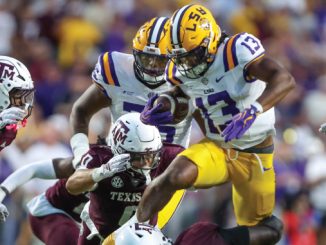 LSU ball carrier in white and yellow jumps over Texas A&M defenders during a crowded college football play.