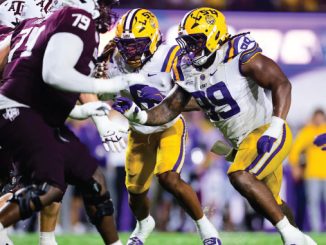 College football players collide on the field during a game, with a LSU player in a white jersey and yellow helmet reaching forward against maroon-clad defenders.