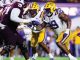 College football players collide on the field during a game, with a LSU player in a white jersey and yellow helmet reaching forward against maroon-clad defenders.