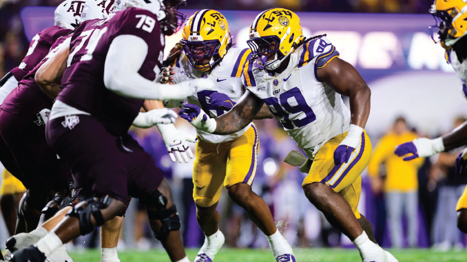 College football players collide on the field during a game, with a LSU player in a white jersey and yellow helmet reaching forward against maroon-clad defenders.