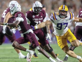 LSU defender wearing number 97 in white and gold chases a Texas A&M quarterback in maroon as he runs with the ball during a college football game.