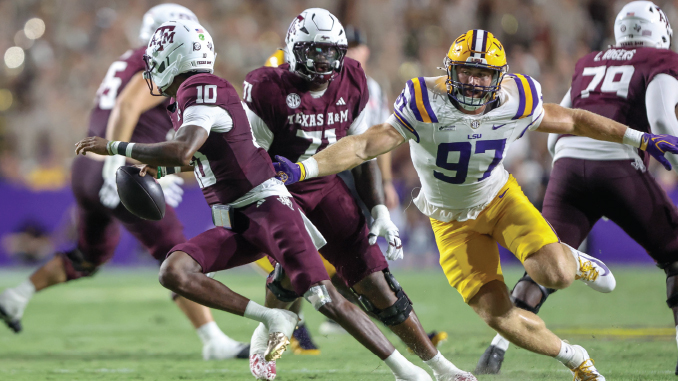 LSU defender wearing number 97 in white and gold chases a Texas A&M quarterback in maroon as he runs with the ball during a college football game.