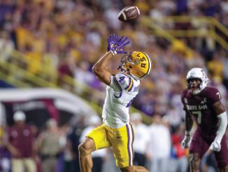 LSU wide receiver in yellow shorts leaps to catch a pass in midair as a defender in maroon pursues in the crowded stadium.