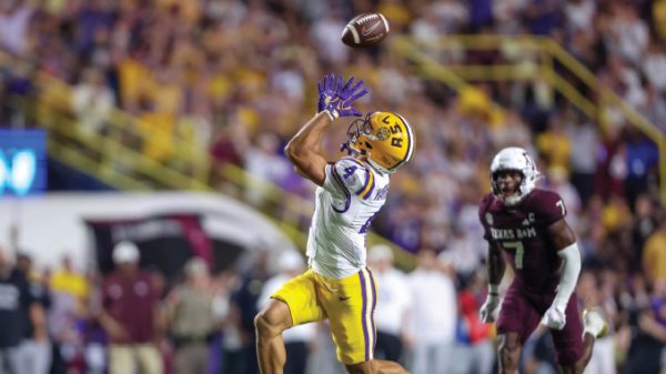 LSU wide receiver in yellow shorts leaps to catch a pass in midair as a defender in maroon pursues in the crowded stadium.