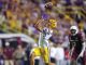LSU wide receiver in yellow shorts leaps to catch a pass in midair as a defender in maroon pursues in the crowded stadium.