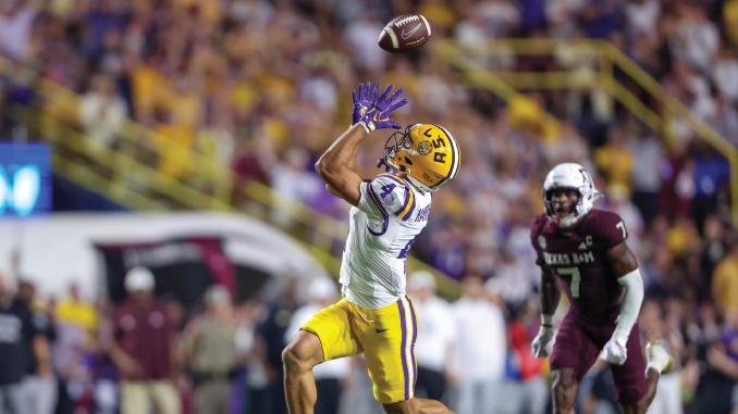 LSU wide receiver in yellow shorts leaps to catch a pass in midair as a defender in maroon pursues in the crowded stadium.