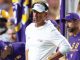 LSU football coach in a white polo and headset standing on the sideline during a game, with players in purple and gold in the background.
