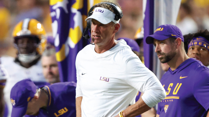 LSU football coach in a white polo and headset standing on the sideline during a game, with players in purple and gold in the background.