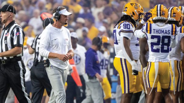 LSU football coach in white attire on the sideline during a game with players in yellow helmets nearby.