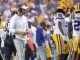 LSU football coach in white attire on the sideline during a game with players in yellow helmets nearby.