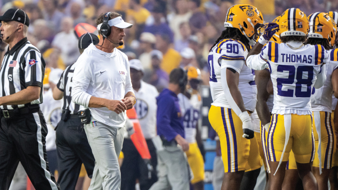 LSU football coach in white attire on the sideline during a game with players in yellow helmets nearby.