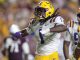 LSU football player in a white jersey and gold pants celebrating on the field during a college game, purple helmet visible.