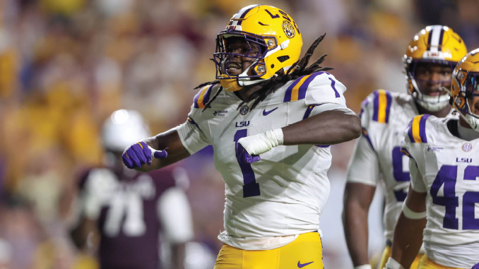 LSU football player in a white jersey and gold pants celebrating on the field during a college game, purple helmet visible.