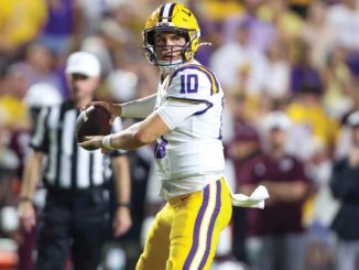 Louisiana State University quarterback in white and gold uniform holding a football as he scans the field during a game.