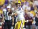 Louisiana State University quarterback in white and gold uniform holding a football as he scans the field during a game.