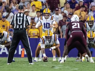 LSU football offense lines up at snap with quarterback #10 under center as defenders prepare behind a maroon-clad opponent; crowded stadium in background.