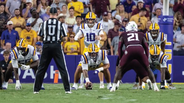 LSU football offense lines up at snap with quarterback #10 under center as defenders prepare behind a maroon-clad opponent; crowded stadium in background.