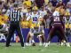 LSU football offense lines up at snap with quarterback #10 under center as defenders prepare behind a maroon-clad opponent; crowded stadium in background.
