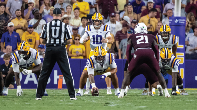 LSU football offense lines up at snap with quarterback #10 under center as defenders prepare behind a maroon-clad opponent; crowded stadium in background.