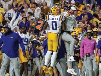 LSU football players celebrate on the sideline after a play; player number 0 Cooley is embraced by a teammate, with a purple-and-gold crowd in the background.