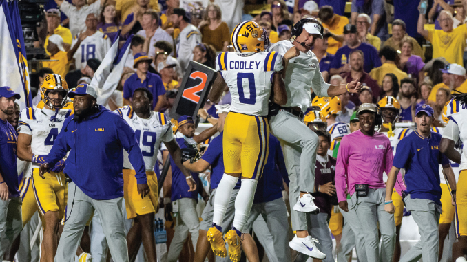 LSU football players celebrate on the sideline after a play; player number 0 Cooley is embraced by a teammate, with a purple-and-gold crowd in the background.