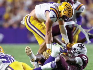 Lineman in LSU yellow pants tackles a Texas A&M player on the ground during a football game.