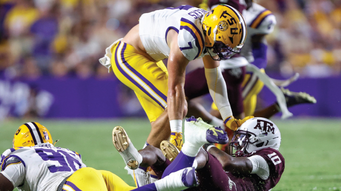 Lineman in LSU yellow pants tackles a Texas A&M player on the ground during a football game.
