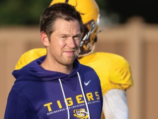 Man in a purple Tigers hoodie on a practice field, with a yellow-jersey player in the background.