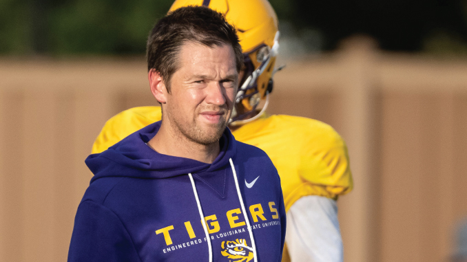 Man in a purple Tigers hoodie on a practice field, with a yellow-jersey player in the background.