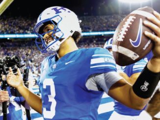 Football quarterback in a blue uniform on a crowded stadium field, ball in hand and ready to throw.