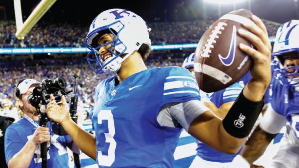 Football quarterback in a blue uniform on a crowded stadium field, ball in hand and ready to throw.