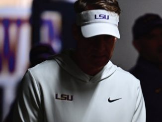 Person wearing a white LSU visor and hoodie with a Nike swoosh, head bowed in a dimly lit setting.