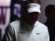 Person wearing a white LSU visor and hoodie with a Nike swoosh, head bowed in a dimly lit setting.