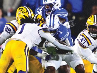 Football game: LSU players (yellow pants) tackle a blue-clad defender near the line of scrimmage during a burst of tackles in progress in a crowded stadium.