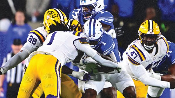 Football game: LSU players (yellow pants) tackle a blue-clad defender near the line of scrimmage during a burst of tackles in progress in a crowded stadium.
