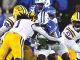 Football game: LSU players (yellow pants) tackle a blue-clad defender near the line of scrimmage during a burst of tackles in progress in a crowded stadium.