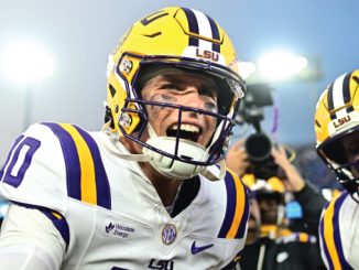 LSU quarterback grins during a celebration with teammates on the field, wearing a yellow helmet with LSU logo.