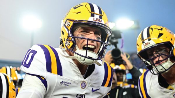 LSU quarterback grins during a celebration with teammates on the field, wearing a yellow helmet with LSU logo.