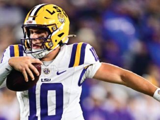 LSU quarterback wearing number 10 runs with a football in a white and purple uniform during a game.