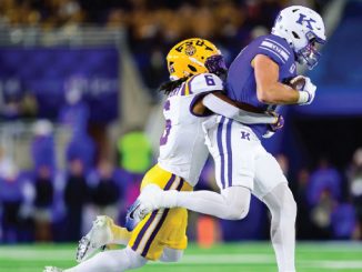 A football player in a purple jersey carries the ball while a white-jersey defender tackles him on a bright field at night stadiums.