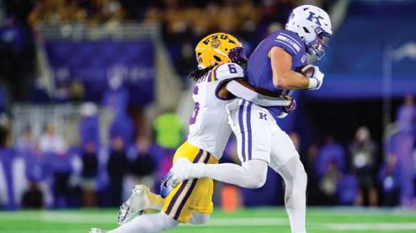 A football player in a purple jersey carries the ball while a white-jersey defender tackles him on a bright field at night stadiums.