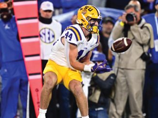 LSU football player wearing #14 in white jersey and yellow pants makes a sideline catch, with photographers and fans watching in the background.